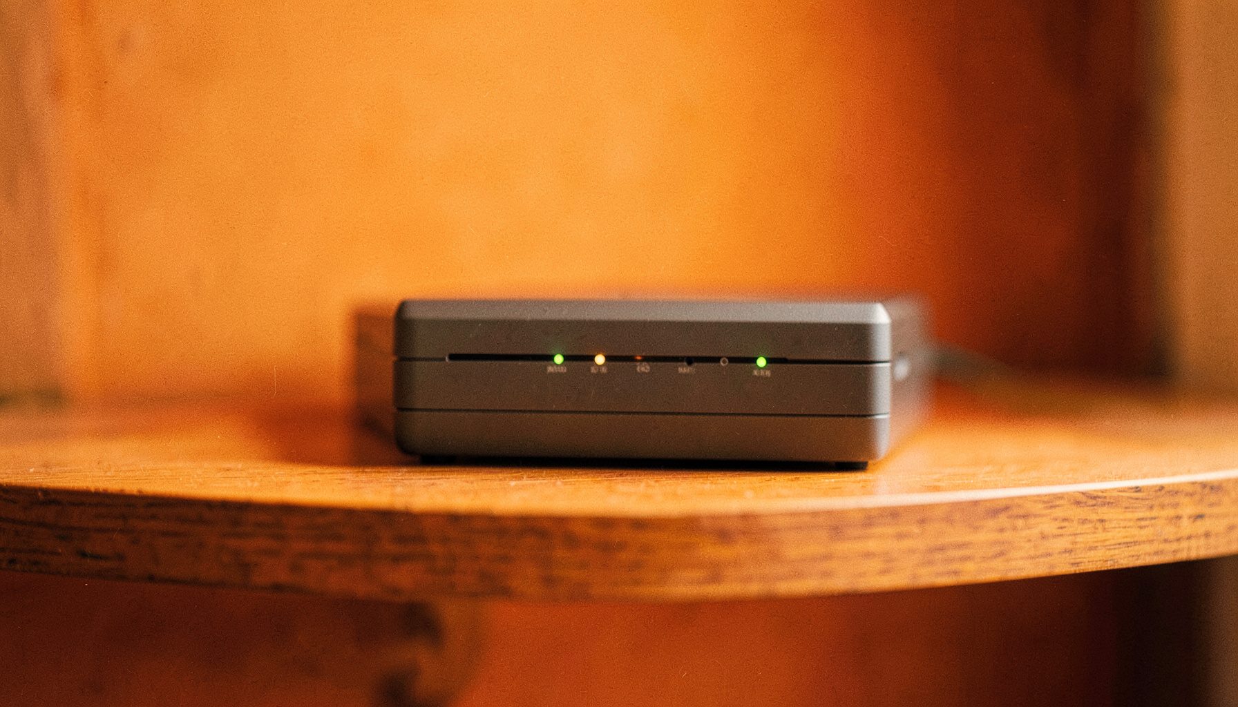 A compact cable modem with indicator lights, placed on a wooden shelf, soft natural light