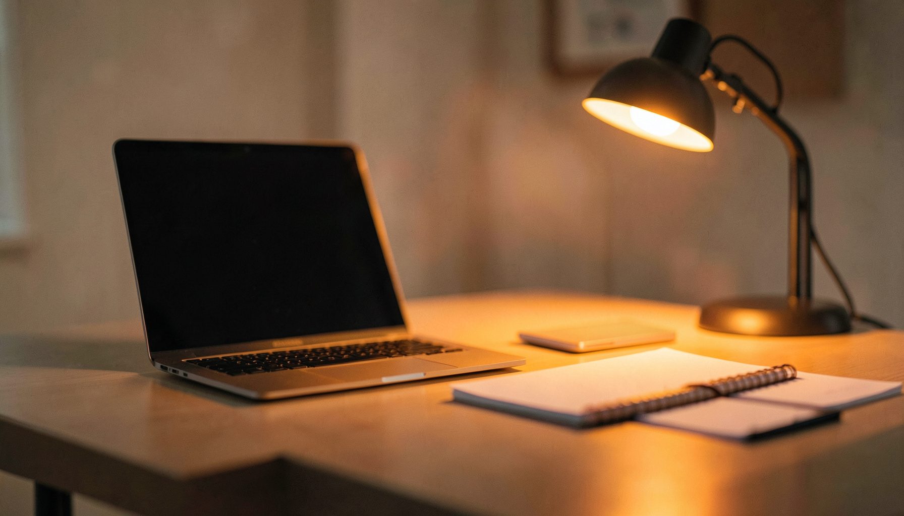 A clean, modern desk workspace with a laptop, notebook, and a warm desk lamp glow against a softly blurred background in neutral tones