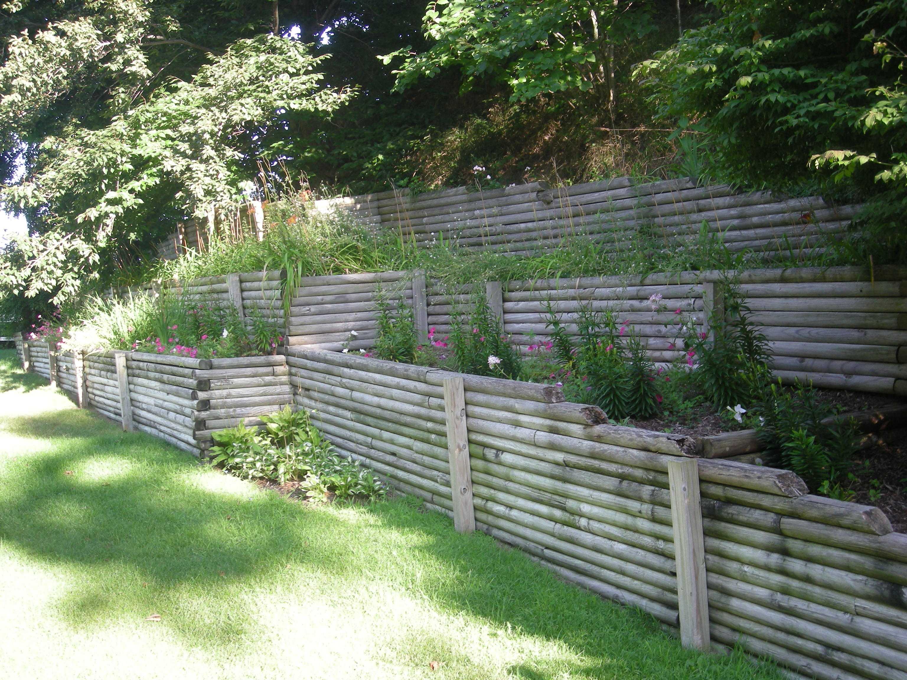 Tiered wooden log retaining wall garden with colorful flowers and greenery, surrounded by trees and a grassy lawn in day