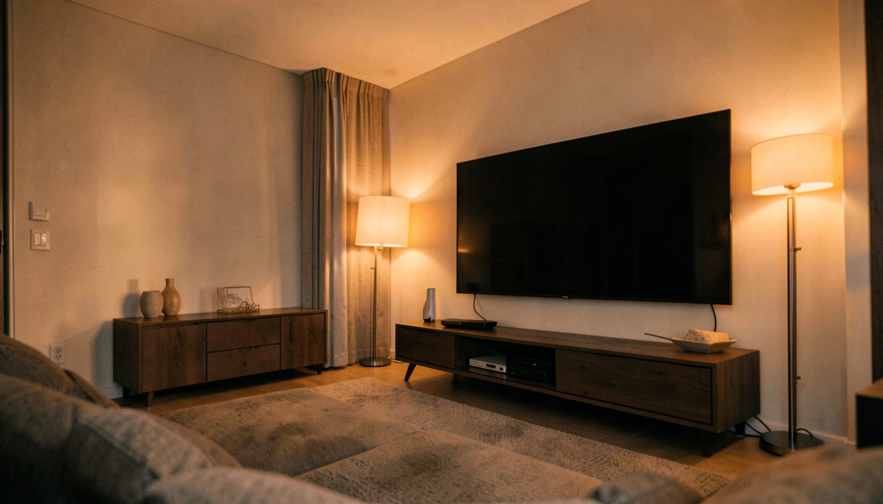 A wide view of a modern living room with a large flat-screen TV, a sleek router on a media console, and soft warm lighting from a floor lamp, in neutral beige and charcoal tones.